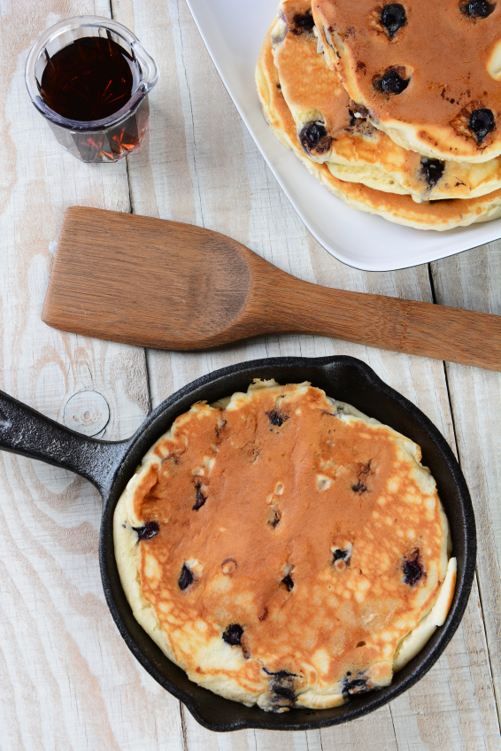 High angle shot of a cast iron skillet with a a freshly made blueberry pancake. A plate with more pancakes, a spatula and syrup pitcher fill the space, on a rustic farmhouse style kitchen table.